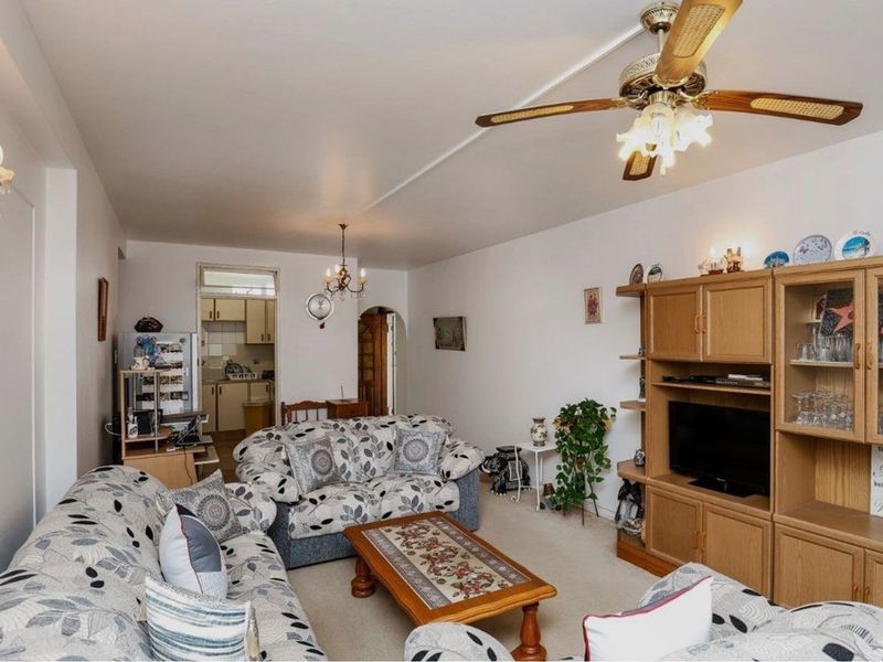 A spacious open-plan living area featuring a ceiling fan, neutral carpeting, and an adjacent kitchen space visible in the background.