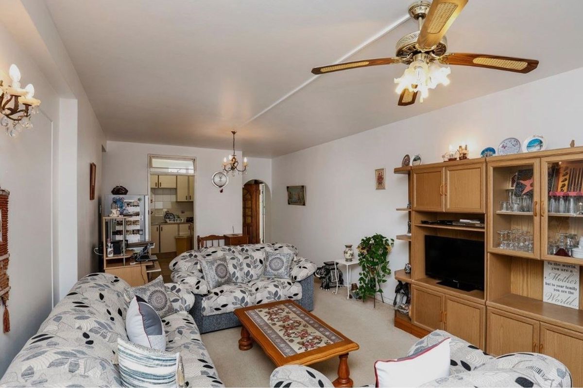 A spacious open-plan living area featuring a ceiling fan, neutral carpeting, and an adjacent kitchen space visible in the background.