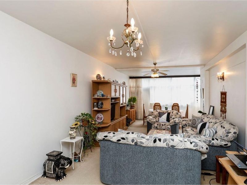 A spacious open-plan living and dining area featuring neutral carpeting, a ceiling fan, and a classic chandelier, offering a versatile layout for daily living.