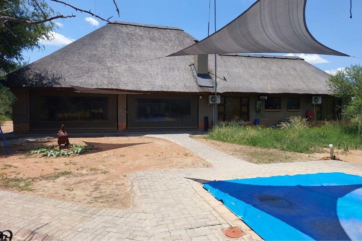 Rear view of a thatched roof house featuring a paved patio area, swimming pool with cover, and shade sail structure.