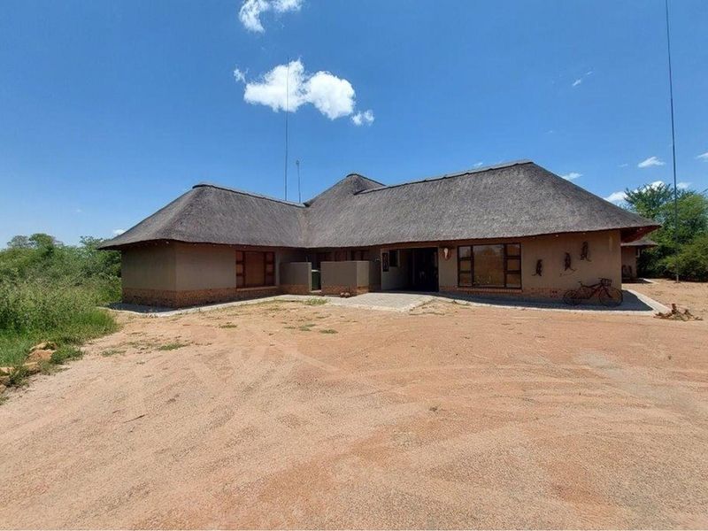 A single-story house featuring traditional thatched roofing, earthy exterior walls, and a large, open sandy driveway area.