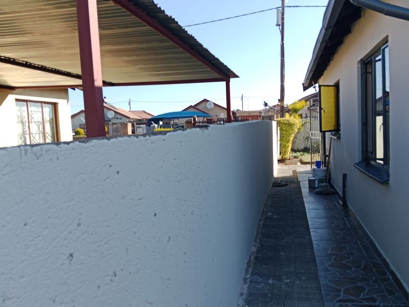 A side exterior view showing a paved walkway, a boundary wall, and a metal carport structure adjacent to the main house.