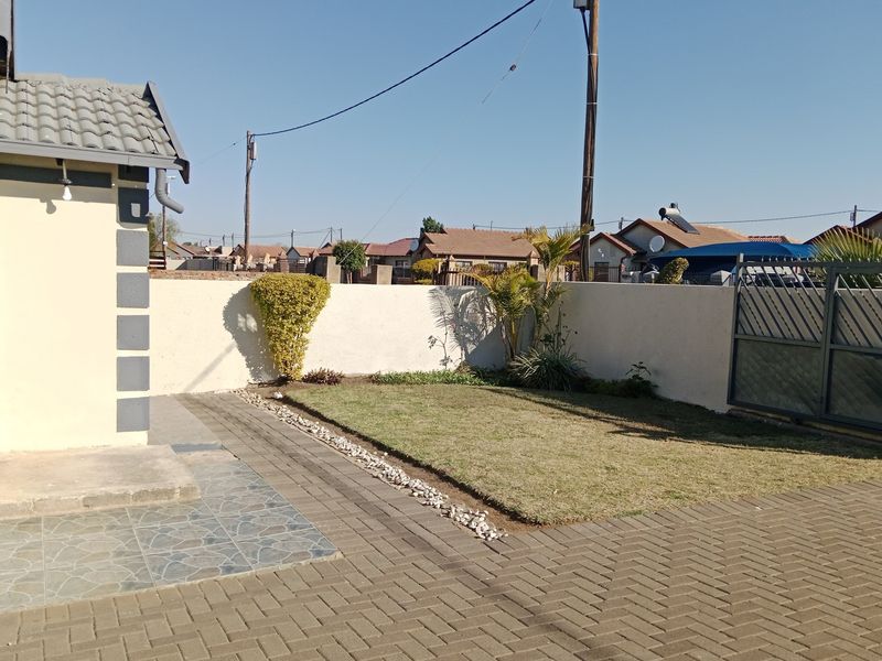 A neat rear exterior area featuring a paved driveway, a small lawn, a tiled patio section, and secure boundary walls with a metal gate.