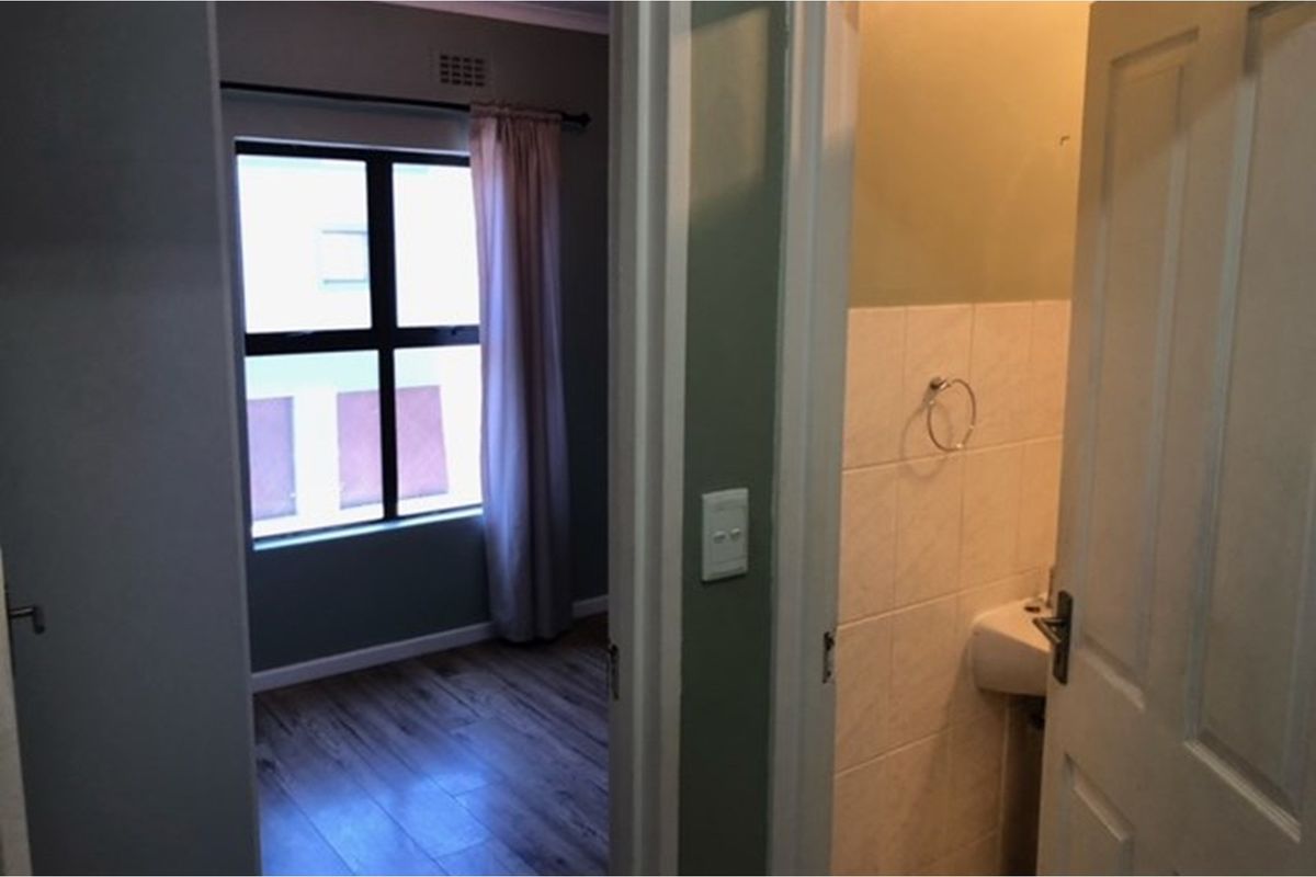 A view through a doorway showing a bedroom with wood-look flooring and a small adjacent bathroom with tiled walls and a wall-mounted basin.