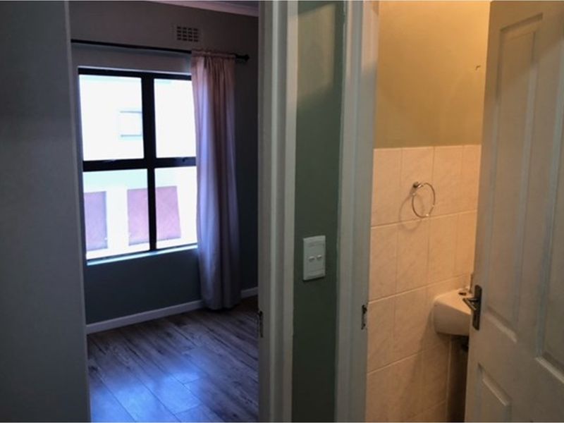 A view through a doorway showing a bedroom with wood-look flooring and a small adjacent bathroom with tiled walls and a wall-mounted basin.