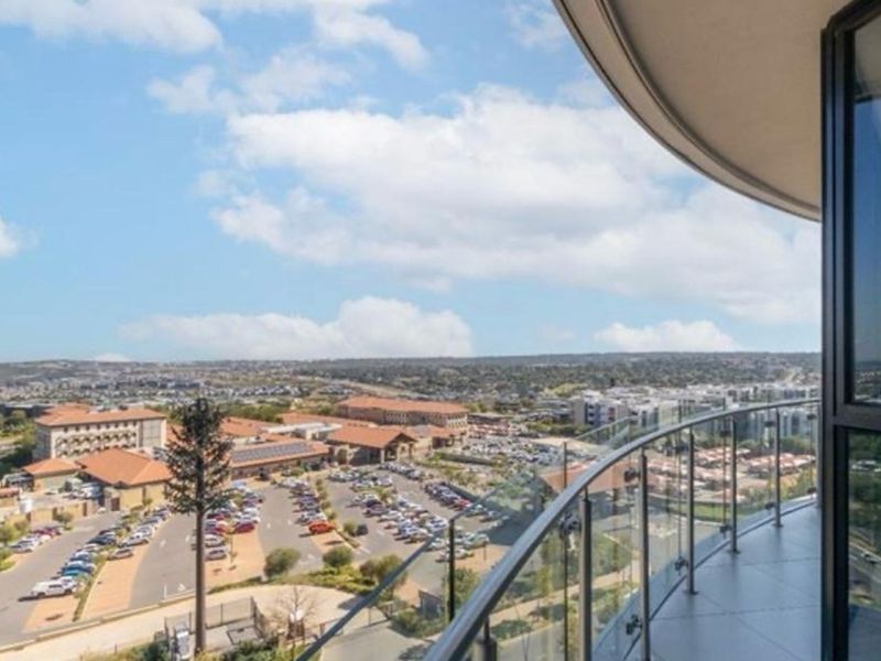 A spacious balcony with a glass balustrade offering expansive elevated views over the surrounding neighbourhood and commercial area.