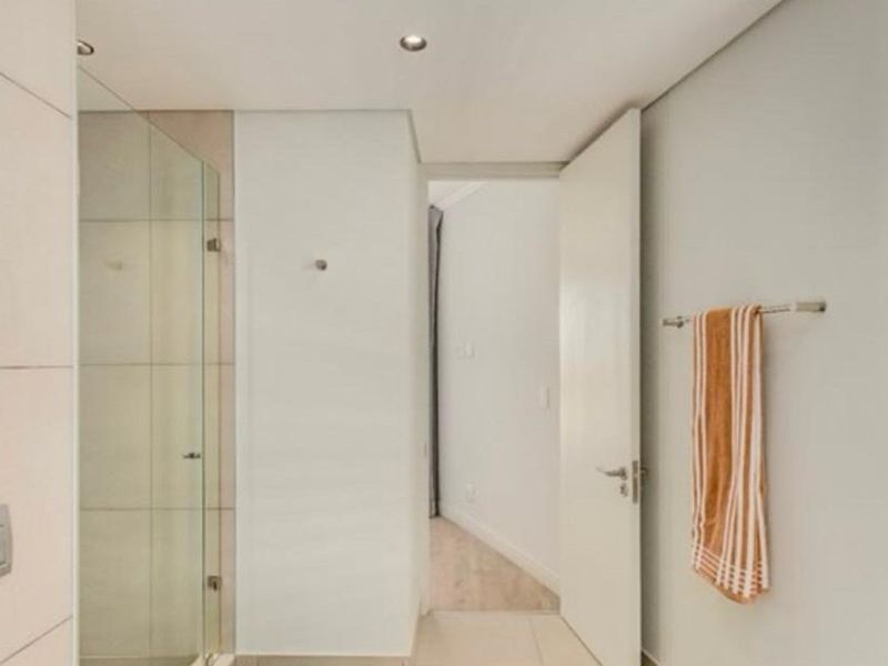 A clean and modern bathroom featuring a glass-enclosed shower, neutral tiled flooring, and recessed lighting, leading into an adjacent room.
