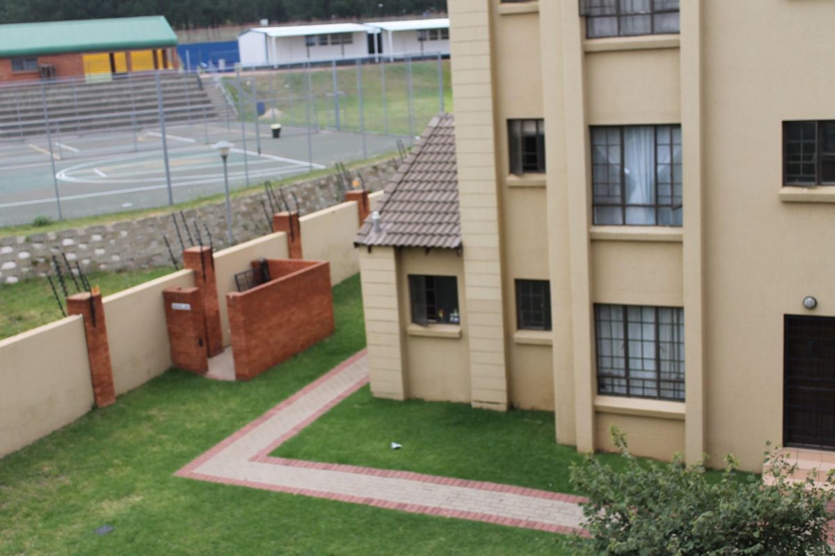 Elevated view of the apartment complex exterior, featuring a paved walkway, landscaped lawn, and perimeter wall with security fencing.
