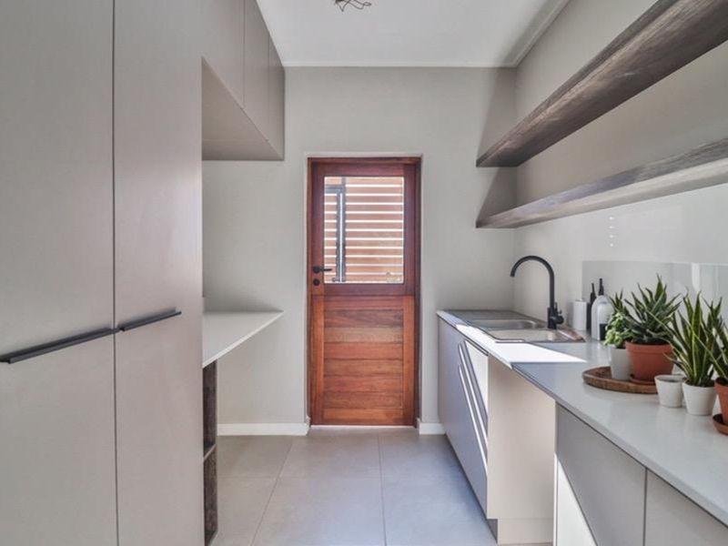 A modern, well-appointed scullery featuring sleek cabinetry, stone countertops, a stainless steel sink, and floating timber shelving with direct exterior access.