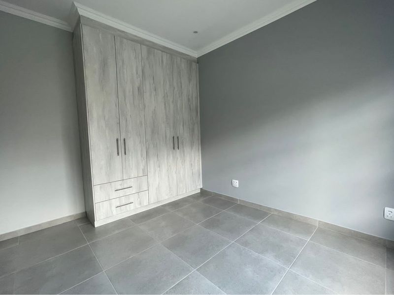 A clean, modern bedroom featuring neutral grey floor tiles, light wood-grain built-in cupboards, and crisp white crown molding.