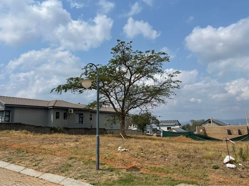 A view of a vacant residential plot in a developed estate, featuring established trees, a street lamp, and surrounding residential properties.