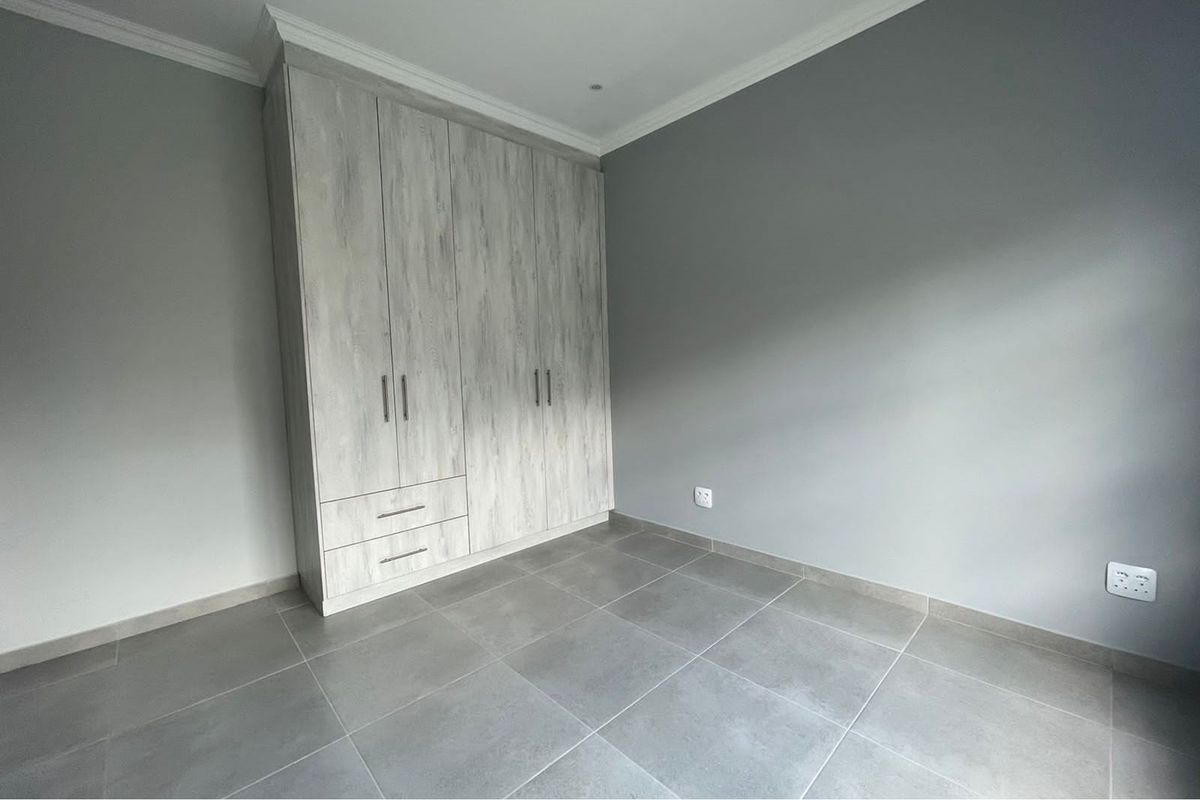 A clean, modern bedroom featuring neutral grey floor tiles, light wood-grain built-in cupboards, and crisp white crown molding.