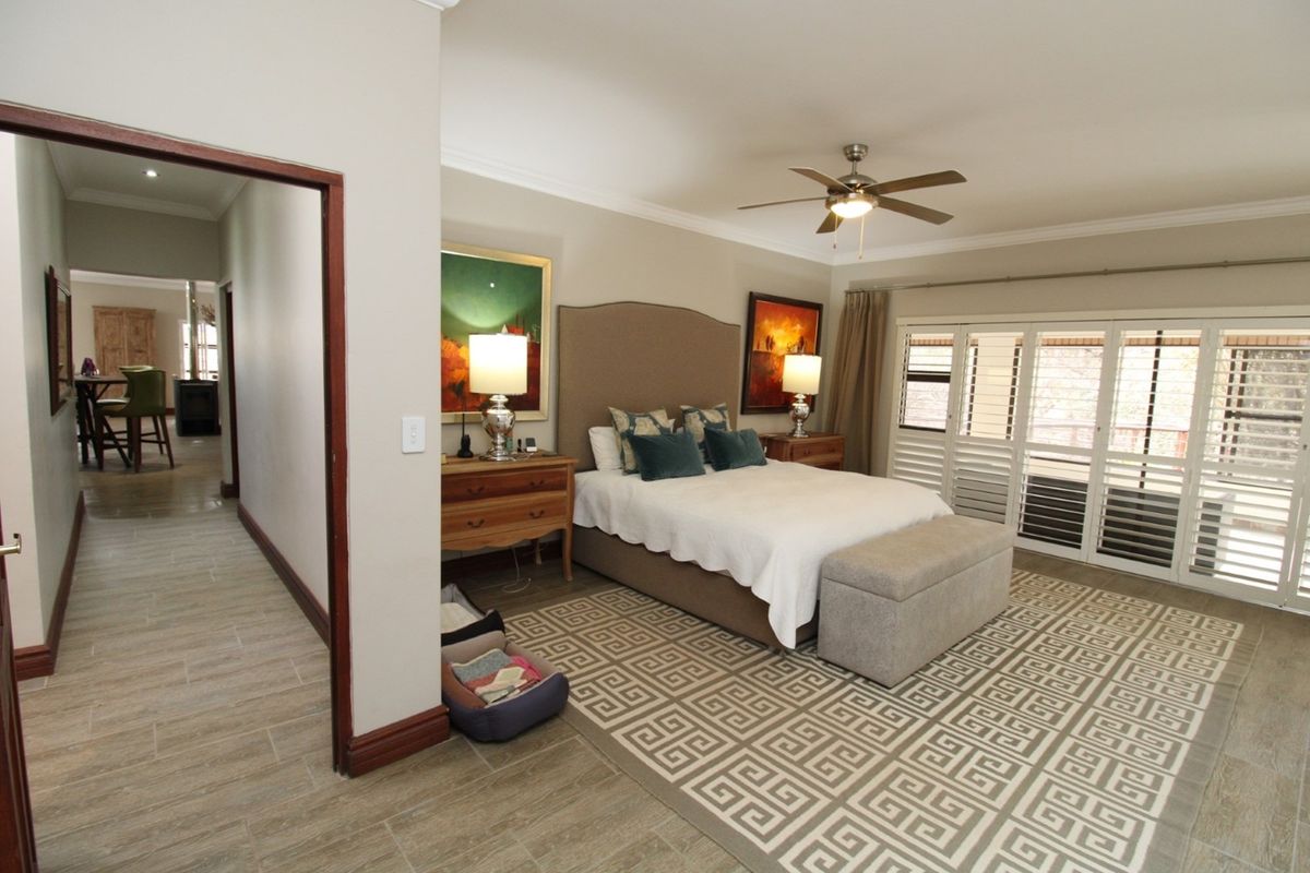Spacious bedroom featuring tiled flooring, a ceiling fan, and plantation shutters leading to an outdoor area, with an adjacent hallway view.