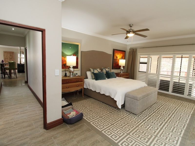 Spacious bedroom featuring tiled flooring, a ceiling fan, and plantation shutters leading to an outdoor area, with an adjacent hallway view.