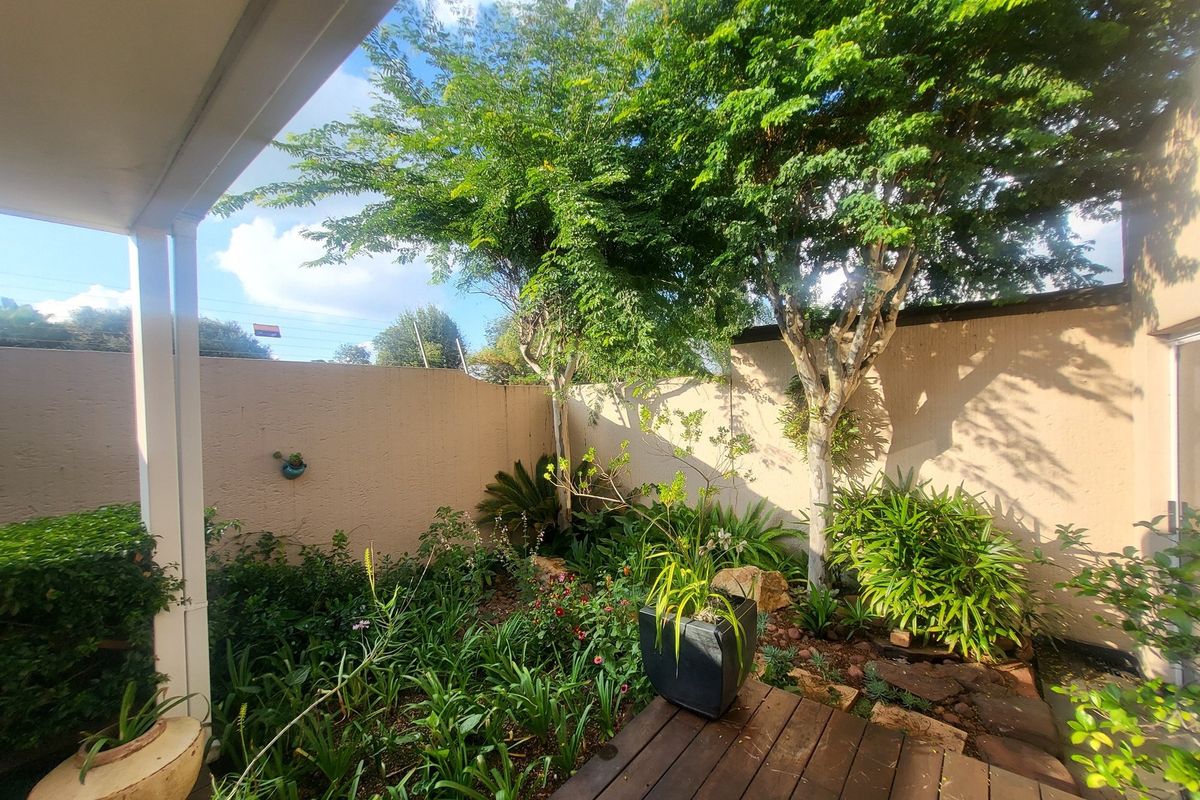 A private rear garden area featuring established trees, landscaped garden beds, and a wooden deck, enclosed by high boundary walls.
