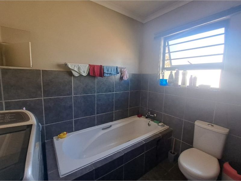 A functional bathroom featuring dark wall tiling, a built-in bathtub, a toilet, and a window with security bars.