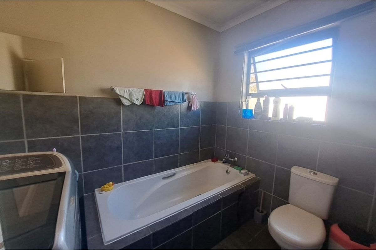 A functional bathroom featuring dark wall tiling, a built-in bathtub, a toilet, and a window with security bars.