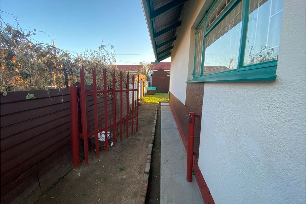 A side exterior view of the property featuring a concrete pathway, metal security fencing, and painted exterior walls with window frames.