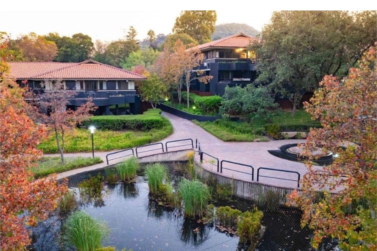 A scenic view of the apartment complex grounds featuring landscaped gardens, a tranquil pond with a bridge, and mature trees.