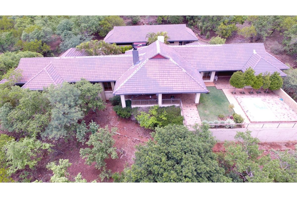 Aerial view of a spacious residential property featuring a tiled roof, covered patio area, swimming pool, and surrounding landscaped garden in a natural setting.