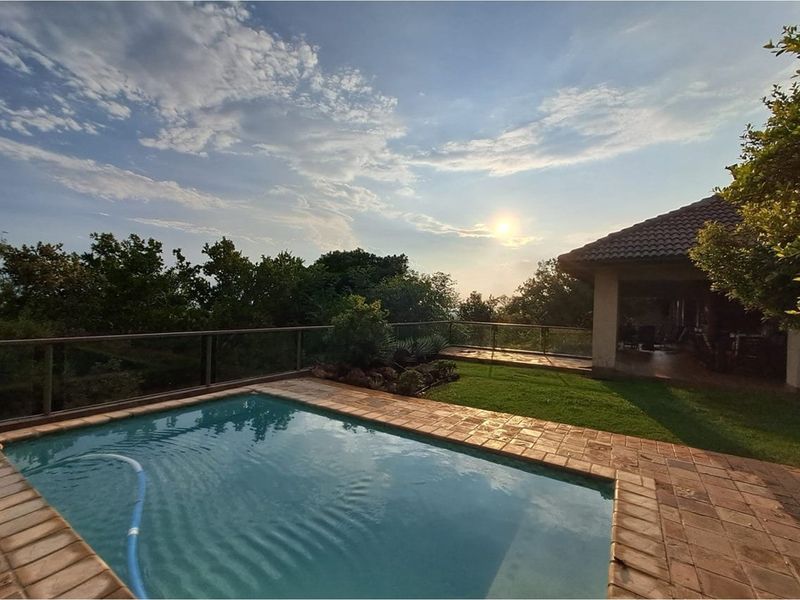 Outdoor pool area featuring a paved surround, glass balustrade, and a covered patio overlooking the garden and surrounding landscape.
