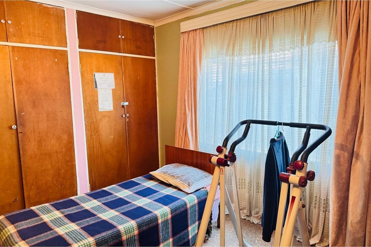A bedroom featuring built-in wooden cupboards and a large window with curtains, offering a functional space with potential for modernization.