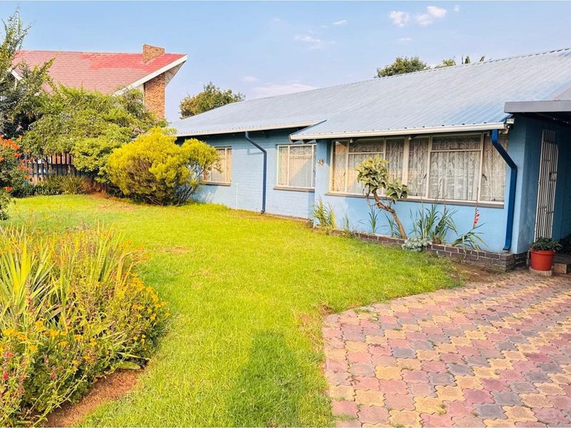 Front view of a single-story house featuring a blue brick facade, corrugated metal roof, and a paved driveway set within a landscaped garden.