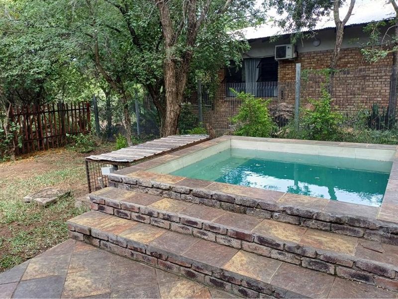 A raised swimming pool featuring stone tile cladding, set within a garden area adjacent to a brick-walled house with an exterior air conditioning unit.