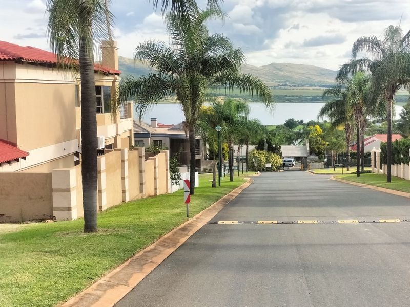 A view looking down a paved residential street within a well-maintained estate, featuring landscaped verges, palm trees, and a scenic lake backdrop.