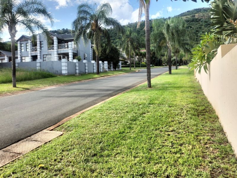 A view of a paved residential street in a well-maintained estate, featuring manicured lawns, palm trees, and surrounding mountain views.