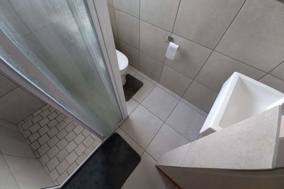 A compact, modern bathroom featuring a glass-enclosed shower, a wall-mounted toilet, and a minimalist basin with neutral floor-to-ceiling tiling.