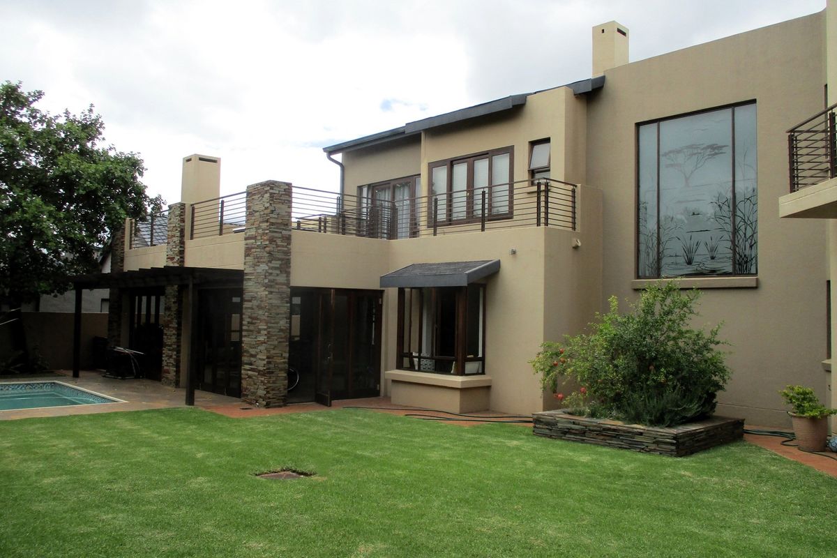 Rear view of a multi-story house featuring a swimming pool, landscaped garden, stone-clad pillars, and a spacious upper-level balcony with metal railings.