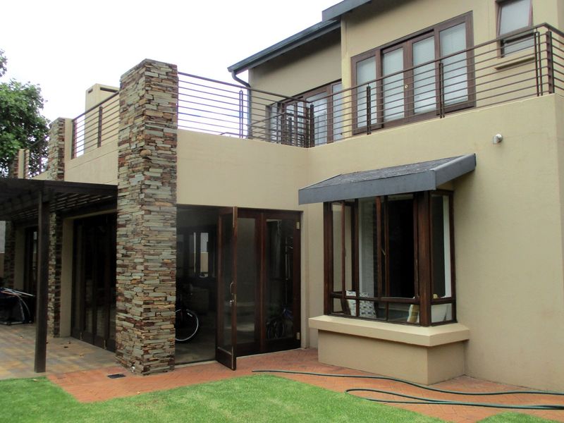 Rear view of a double-story house featuring a stone-clad pillar, a balcony with metal railings, and a paved patio area leading to the garden.