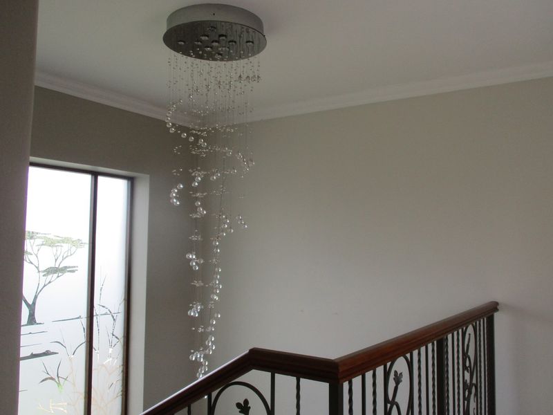A bright hallway featuring a decorative crystal chandelier, a wooden handrail with metal balustrades, and a tall window with frosted glass detailing.