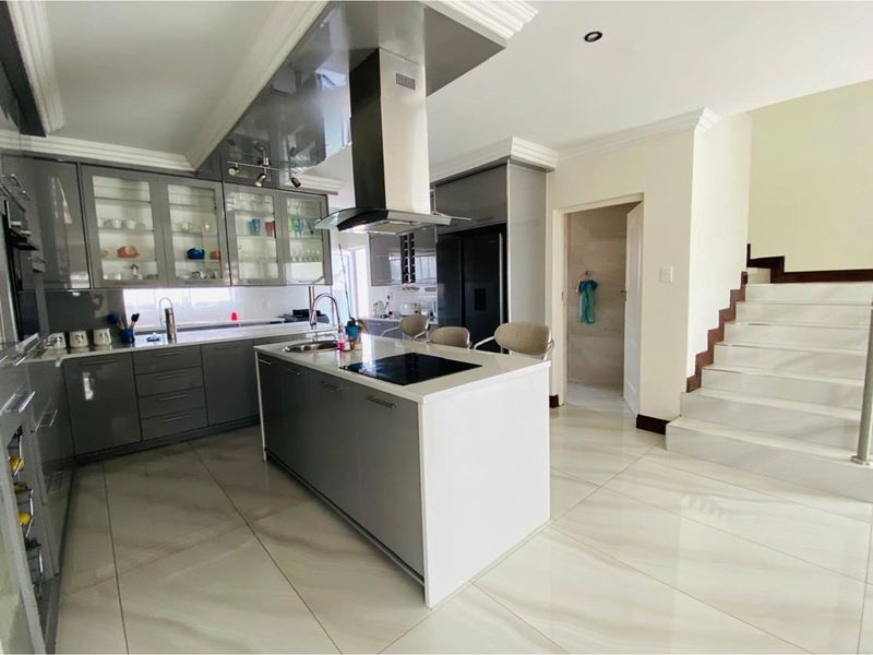 Modern kitchen featuring high-gloss cabinetry, a central island with a built-in cooktop, stainless steel extractor fan, and tiled flooring, adjacent to a staircase.