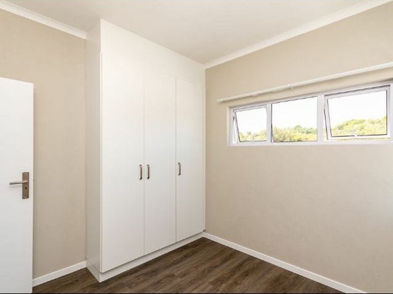 A clean, empty bedroom featuring modern laminate flooring, built-in white cupboards, and top-hung windows with a curtain rail.