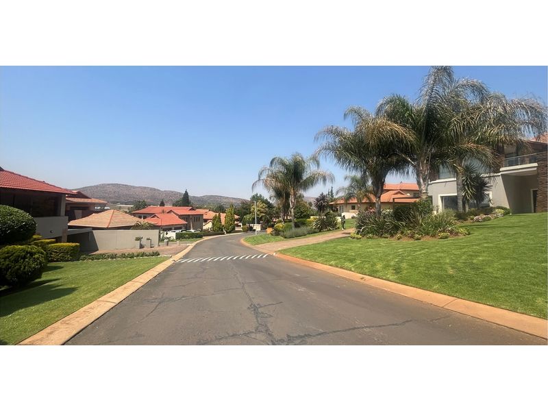 A view of a paved residential street within a landscaped estate, featuring manicured lawns, mature palm trees, and surrounding residential properties.