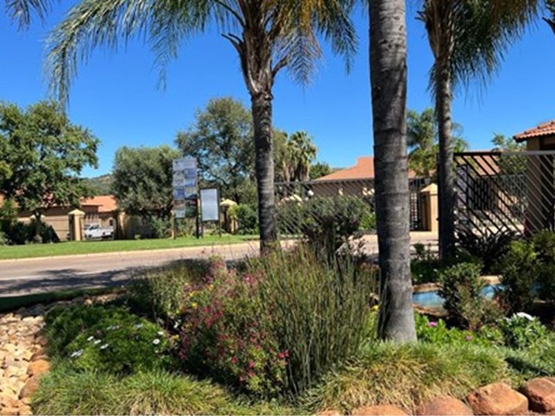 A view of the residential estate entrance featuring landscaped gardens, mature palm trees, and secure perimeter fencing.