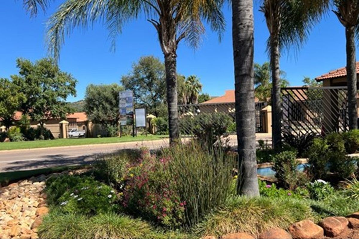 A view of the residential estate entrance featuring landscaped gardens, mature palm trees, and secure perimeter fencing.