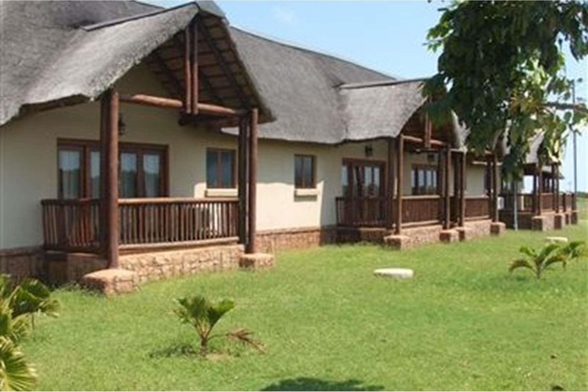 A view of the rear exterior of a farm-style building featuring a thatched roof, timber support beams, and covered patio areas overlooking a lawn.