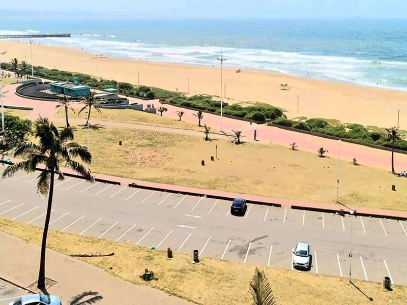 Elevated view overlooking the coastal promenade, public parking area, and sandy beach with ocean waves.