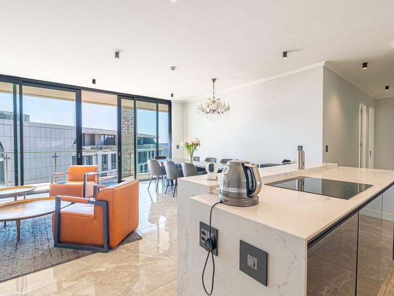 Modern open-plan living area featuring a sleek stone kitchen island with integrated hob, marble-look flooring, and large sliding doors leading to a balcony.
