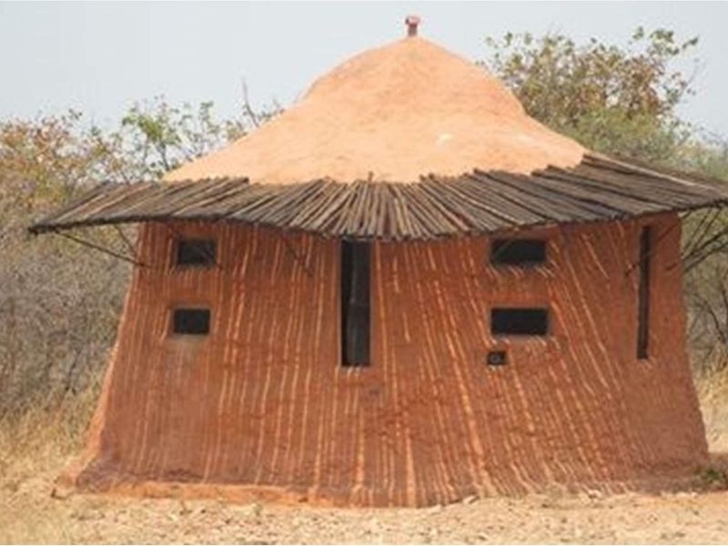 A traditional-style round hut structure with a thatched roof and mud-plastered walls, situated in a rural farm landscape.