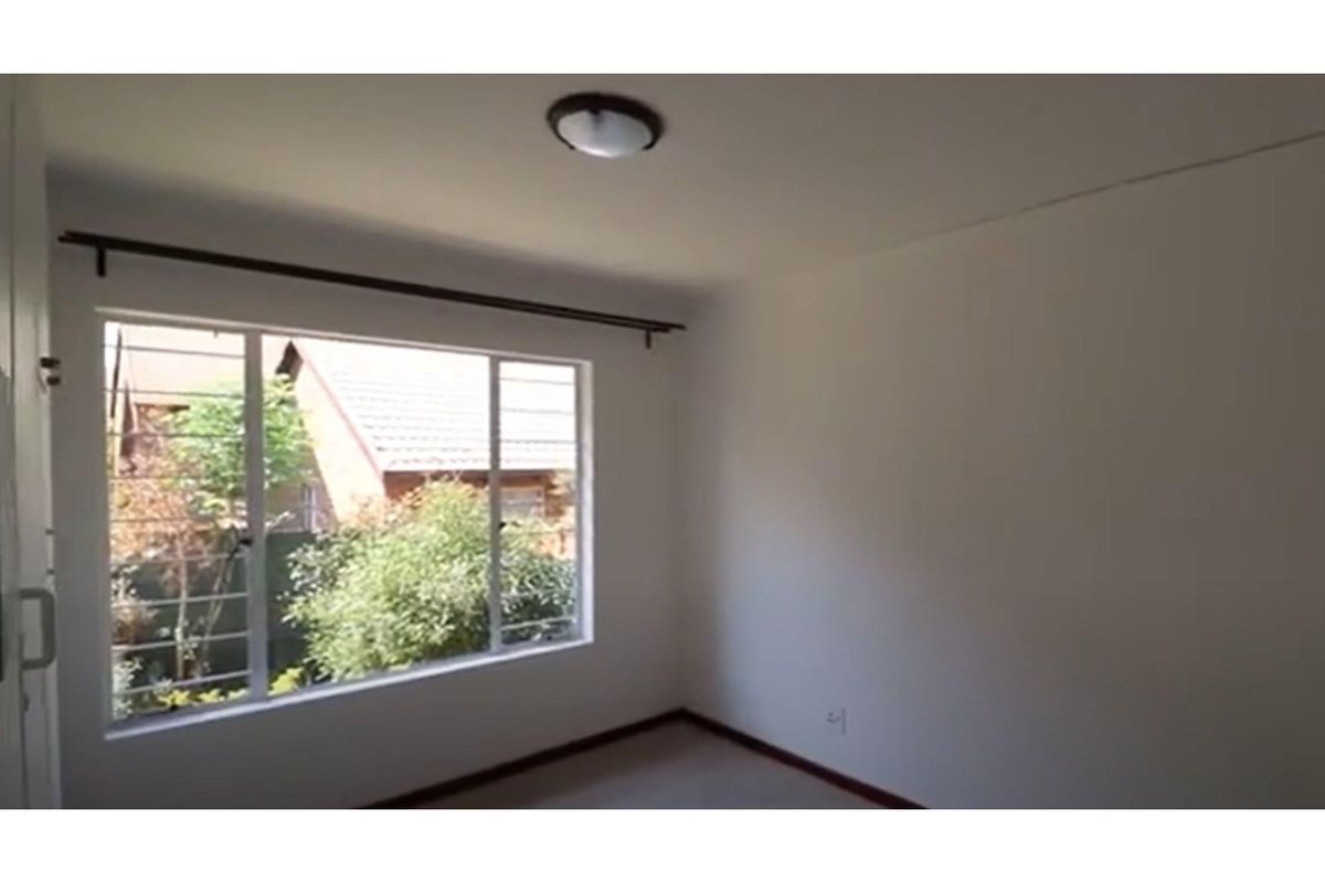 A bright, empty bedroom featuring a large window with security bars, neutral walls, and a simple ceiling light fixture.