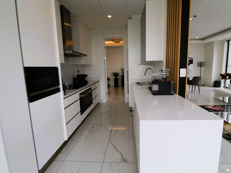 Modern galley-style kitchen featuring sleek white cabinetry, stone countertops, integrated oven, built-in microwave, and a stainless steel extractor fan with polished marble-look floor tiles.