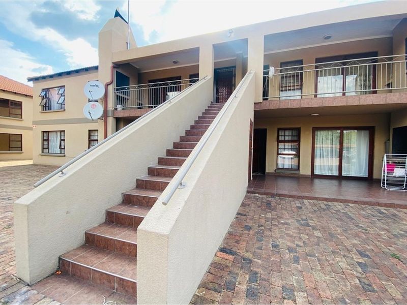 Exterior view of a townhouse complex featuring a central staircase leading to an upper balcony, paved courtyard, and multiple access points.