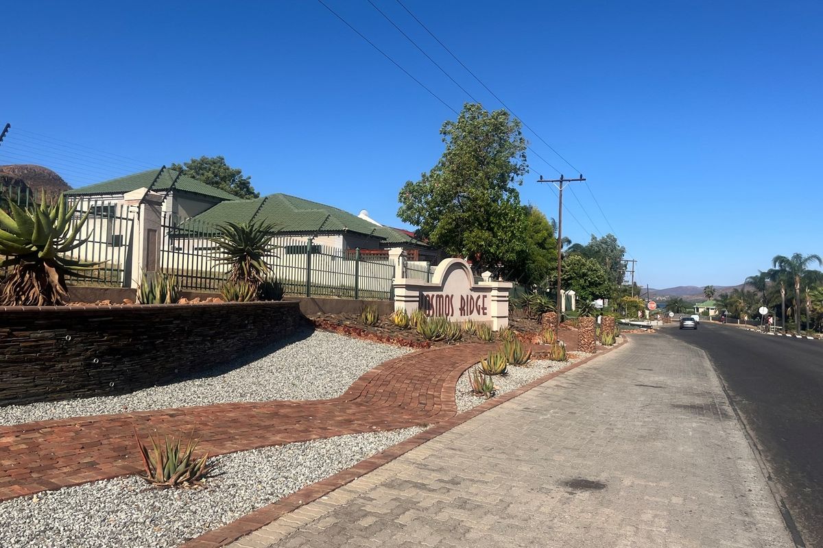 Street view of the Cosmos Ridge estate entrance, featuring a paved walkway, landscaped garden beds with aloes, and a stone-clad retaining wall.