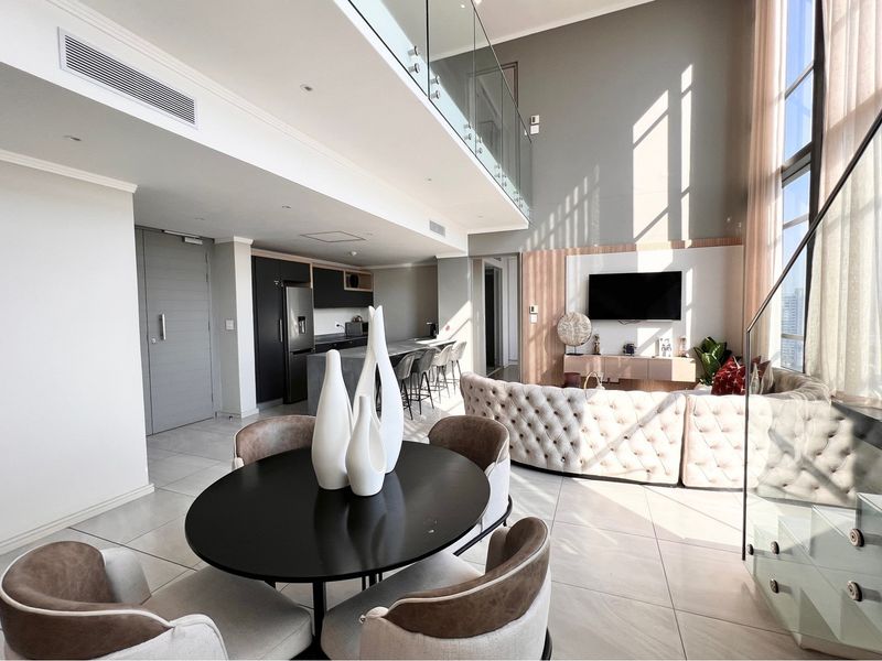 Spacious open-plan penthouse living area featuring double-height ceilings, modern kitchen with island, and a glass-railed mezzanine level with high-end finishes.