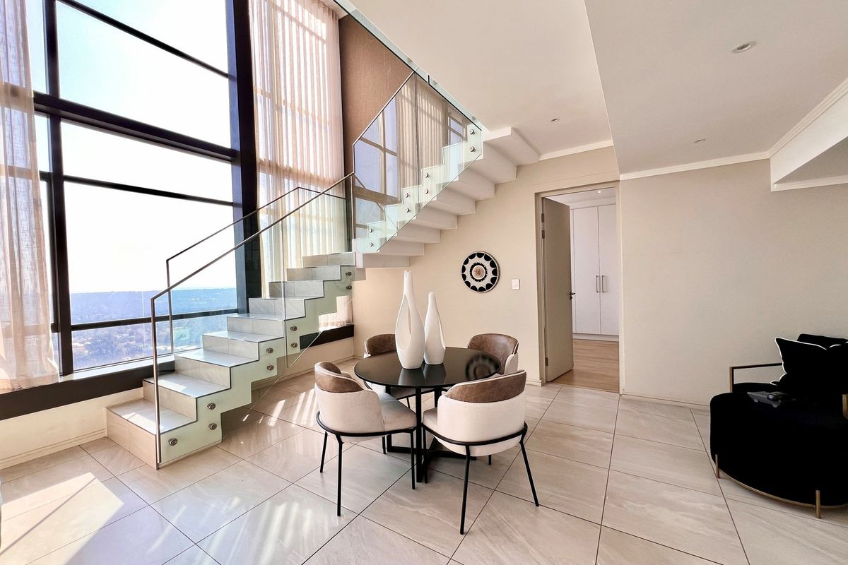 Modern living area featuring a striking glass staircase, floor-to-ceiling windows with expansive views, and sleek tiled flooring in a contemporary penthouse setting.