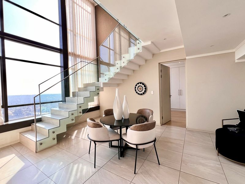 Modern living area featuring a striking glass staircase, floor-to-ceiling windows with expansive views, and sleek tiled flooring in a contemporary penthouse setting.